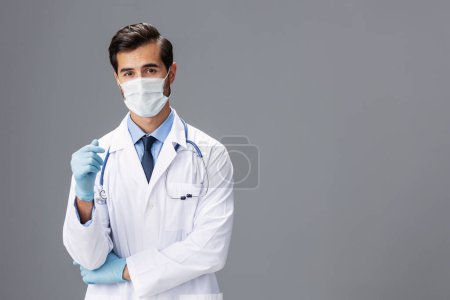 Téléchargez les photos : Un médecin masculin en manteau blanc et masque médical et gants stériles regarde la caméra sur un fond gris isolé, espace de copie, espace pour le texte. Photo de haute qualité - en image libre de droit