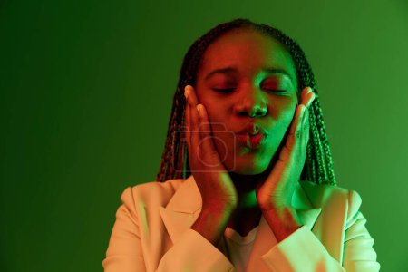 Téléchargez les photos : Afro-américaine femme portrait entreprise sourire sur fond vert dans la lumière au néon, couleur mixte lumière, couleurs vives. Beauté femme afro-américaine avec des émotions pigtails. Photo de haute qualité - en image libre de droit