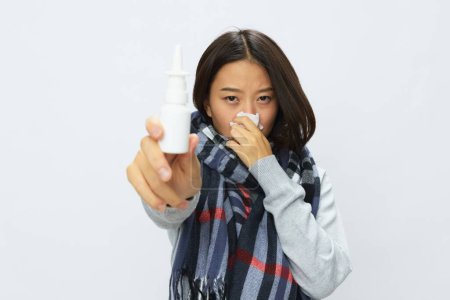 Foto de La mujer asiática resfriada de gripe con nariz tapada sostiene el aerosol nasal de alergia con dolor de garganta en bufanda a cuadros sobre fondo blanco. Foto de alta calidad - Imagen libre de derechos