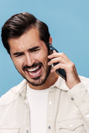 Foto de Retrato de la rabia morena masculina hablando por teléfono en primer plano, descontenta, sobre un fondo azul en una camiseta blanca y jeans, espacio para copiar. Foto de alta calidad - Imagen libre de derechos