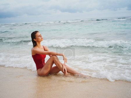 Téléchargez les photos : Une jeune femme en maillot de bain rouge est assise sur le sable avec un beau bronzage et regarde l'océan sous les tropiques sur une plage insulaire. Photo de haute qualité - en image libre de droit