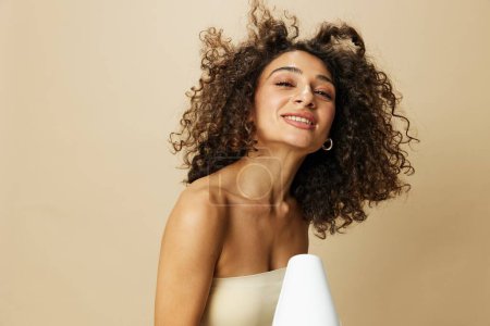 Photo for Woman dries curly hair with blow dryer, home beats styling products, smile on beige background. High quality photo - Royalty Free Image