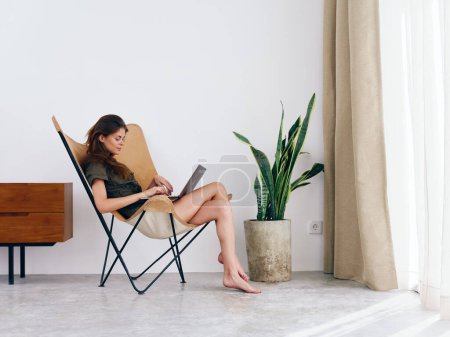 Téléchargez les photos : Femme assise sur une chaise avec un ordinateur portable regardant un film et les médias sociaux à la maison sourire, style de vie scandinave intérieur élégant moderne, espace de copie. Photo de haute qualité - en image libre de droit