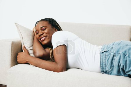 Téléchargez les photos : Heureuse femme afro-américaine dormant à la maison sur le canapé, sourire de repos et de vacances, fond clair, sommeil de jour, dépression de fatigue. Style de vie détendu à la maison, jour de congé. Photo de haute qualité - en image libre de droit
