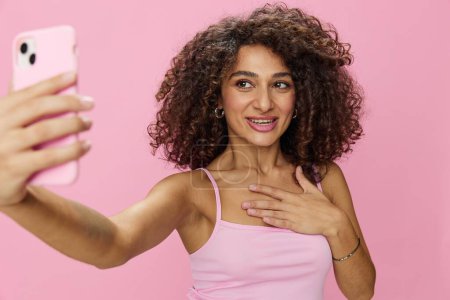 Foto de La bloguera que sostiene una videollamada telefónica se toma selfies, con el pelo rizado en una camiseta de sonrisa rosa y jeans posando sobre un fondo rosa, espacio para copiar, tecnología y redes sociales, en línea. Foto de alta calidad - Imagen libre de derechos