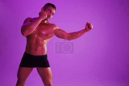Téléchargez les photos : Homme bodybuilder boxer entraînement musculaire avec torse nu. Publicité, sport, mode de vie actif, lumière violette colorée, compétition, concept de défi. . Photo de haute qualité - en image libre de droit