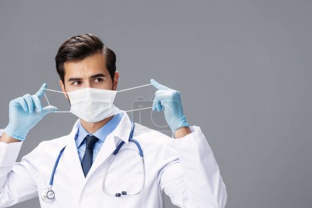 Photo for A male doctor in a white coat and medical mask and sterile gloves looks at the camera on a gray isolated background, copy space, space for text. High quality photo - Royalty Free Image