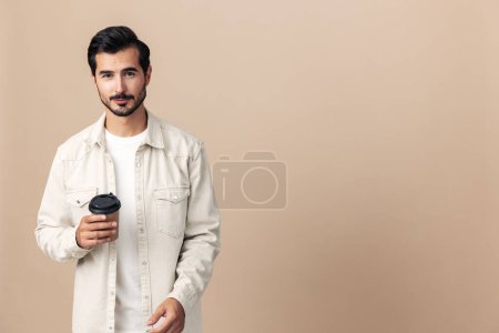 Foto de Retrato de una elegante sonrisa de hombre con una taza de café para ir a burlarse de un fondo beige en una camiseta blanca, estilo de ropa de moda, espacio. Foto de alta calidad - Imagen libre de derechos