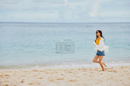 Foto de Mujer deportiva corre a lo largo de la playa en ropa de verano en la arena en una camiseta amarilla y pantalones cortos de mezclilla camisa blanca volando vista al mar pelo. Foto de alta calidad - Imagen libre de derechos