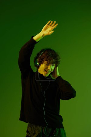Photo for Man wearing headphones listening to music, dancing and singing, DJ teenager happiness and smile, hipster lifestyle, portrait green background mixed neon light, copy space. High quality photo - Royalty Free Image