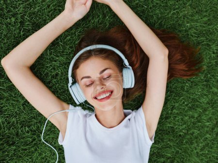 Téléchargez les photos : Femme portant un casque couché sur l'herbe verte dans un T-shirt et écoutant de la musique avec un sourire avec des dents bonheur vacances d'été dans la nature dans le parc. Photo de haute qualité - en image libre de droit