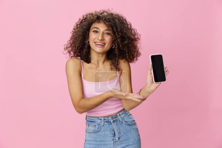 Photo for Woman blogger holding phone video call takes selfies, with curly hair in pink smile t-shirt and jeans poses on pink background, copy space, technology and social media, online. High quality photo - Royalty Free Image
