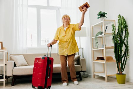 Téléchargez les photos : Heureuse femme âgée avec passeport et billet de voyage emballé une valise rouge, vacances et soins de santé. Une vieille femme souriante se tient joyeusement dans la maison avant le voyage. Photo de haute qualité - en image libre de droit