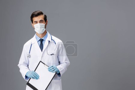 Téléchargez les photos : Un médecin masculin en manteau blanc et un masque médical avec un bloc-notes à la main regarde la caméra sur un fond gris isolé, espace de copie, espace pour le texte. Photo de haute qualité - en image libre de droit