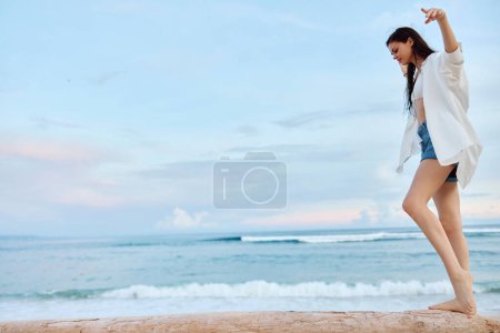 Téléchargez les photos : Joyeux bronzé femme en maillot de bain blanc chemise et short en denim marche sur la plage sur une bûche sur le sable près de l'océan avec les cheveux mouillés après la baignade, la lumière du coucher de soleil à Bali. Photo de haute qualité - en image libre de droit