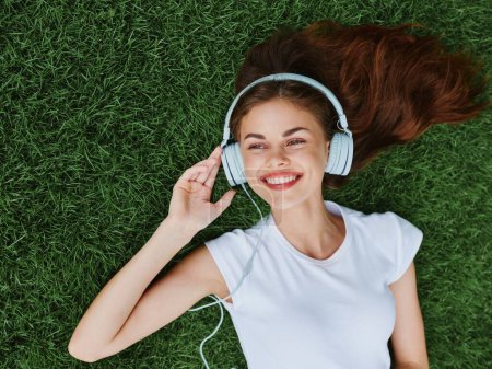 Téléchargez les photos : Une belle femme portant un casque écoute de la musique couchée sur l'herbe dans le parc au printemps et sourit. Photo de haute qualité - en image libre de droit