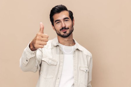 Photo for Portrait of a stylish man smile shows a class sign thumb up on a beige background in a white t-shirt, fashionable clothing style, copy space. High quality photo - Royalty Free Image