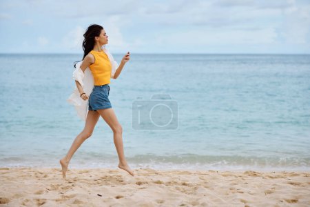Photo for Sports woman runs along the beach in summer clothes on the sand in a yellow tank top and denim shorts white shirt flying hair ocean view, beach vacation and travel. High quality photo - Royalty Free Image