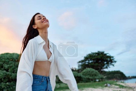 Photo for Brunette woman with long hair in a white shirt and jean shorts tan body abs and happiness fun smile with teeth on the beach and palm trees, vacation summer trip sunset sky. High quality photo - Royalty Free Image