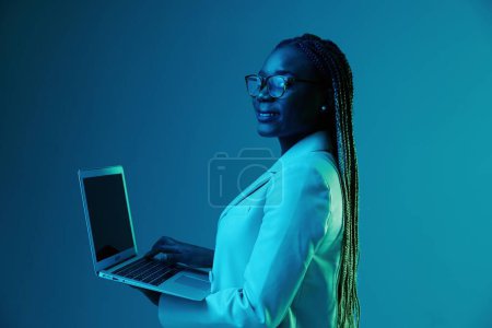 Téléchargez les photos : Africaine femme africaine avec ordinateur portable dans des lunettes sur un fond bleu dans la lumière au néon, lumière mixte de couleur. Stockage de données cybersécurité, pirate informatique, Big Data. Photo de haute qualité - en image libre de droit