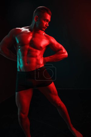 Téléchargez les photos : Homme bodybuilder athlète avec torse nu pose de mode sur fond noir isolé avec lumière colorée néon rouge et vert. Le concept d'un sport sain du corps masculin. Photo de haute qualité - en image libre de droit