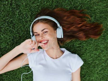 Photo for Woman wearing headphones lying on the green grass in a T-shirt and listening to music with a smile with teeth happiness summer vacations in nature in the park . High quality photo - Royalty Free Image