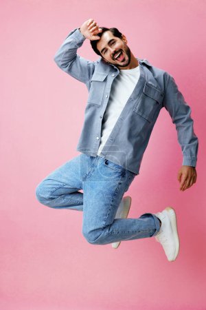 Téléchargez les photos : Homme de mode avec barbe sautant sur fond rose en t-shirt blanc et jean, sourire et émotion joyeuse sur son visage, pleine longueur, espace de copie. Photo de haute qualité - en image libre de droit