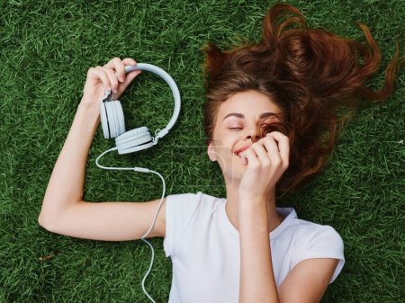 Téléchargez les photos : Une femme allongée sur l'herbe verte avec des écouteurs à la main étendant ses cheveux sur le sol en été, un sourire avec des dents. Photo de haute qualité - en image libre de droit