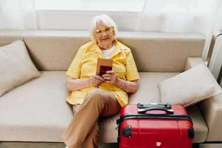 Photo for Happy senior woman with passport and travel ticket packed a red suitcase, vacation and health care. Smiling old woman happily sits on the sofa before the trip. High quality photo - Royalty Free Image