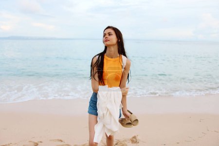 Téléchargez les photos : Une jeune femme après avoir nagé dans l'océan avec un sac à dos en vêtements humides se promène le long de la plage, vacances d'été sur une île au bord de l'océan à Bali coucher de soleil. Photo de haute qualité - en image libre de droit