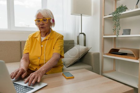 Téléchargez les photos : Heureuse femme âgée avec un ordinateur portable tapant dans un casque sourire assis à la maison sur le canapé dans une chemise jaune, intérieur moderne lumineux, mode de vie communication en ligne. Photo de haute qualité - en image libre de droit
