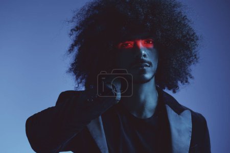 Téléchargez les photos : Portrait de mode d'un homme aux cheveux bouclés sur fond bleu avec une bande rouge de lumière, lumière multicolore, concept tendance et moderne. Photo de haute qualité - en image libre de droit