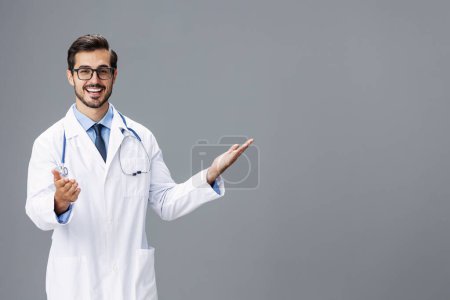 Foto de Hombre sonrisa médico en una bata blanca y gafas para la visión y un estetoscopio mira a la cámara con las manos abiertas sobre un fondo gris aislado, espacio de copia, espacio para el texto, la salud. Foto de alta calidad - Imagen libre de derechos