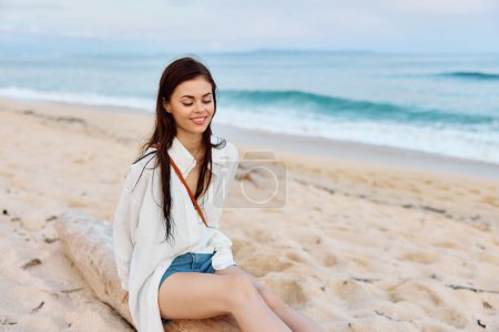Foto de Mujer bronceada feliz sonriendo con dientes en traje de baño blanco camisa y pantalones cortos de mezclilla se sienta en la arena junto al océano con el pelo mojado después de nadar, la luz del atardecer y las nubes de color rosa en Bali. Foto de alta calidad - Imagen libre de derechos
