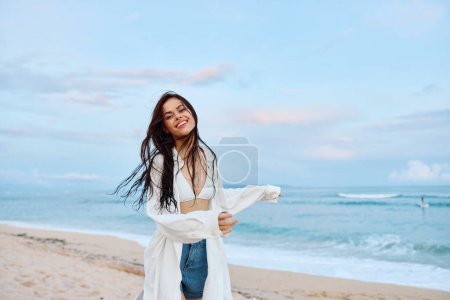 Téléchargez les photos : Brunette femme aux cheveux longs en chemise blanche et short en denim sourire et bonheur en cours d'exécution sur la plage et avoir le sourire amusant avec des dents en face de l'océan, vacances voyage d'été. Photo de haute qualité - en image libre de droit