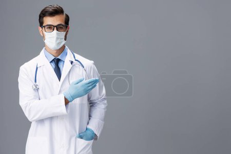 Téléchargez les photos : Médecin masculin vêtu d'un manteau blanc, d'un masque médical et de gants stériles, les lunettes regardent la caméra sur un fond gris isolé, espace de copie, espace pour le texte, santé. Photo de haute qualité - en image libre de droit