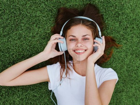 Foto de Una mujer feliz tumbada en el césped verde con una camiseta blanca y escuchando música con sus auriculares sonriendo con las manos en alto. Foto de alta calidad - Imagen libre de derechos