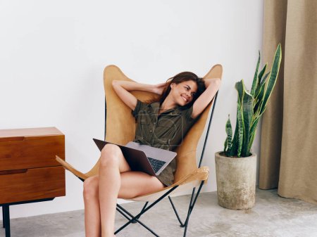 Téléchargez les photos : Femme étudiante indépendante assise sur une chaise avec un travail d'ordinateur portable à la maison sourire et se détendre, style de vie scandinave intérieur élégant moderne, espace de copie. Photo de haute qualité - en image libre de droit