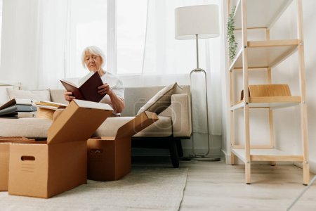 Photo for Elderly woman sits on a sofa at home with boxes. collecting things with memories albums with photos and photo frames moving to a new place cleaning things and a happy smile. High quality photo - Royalty Free Image