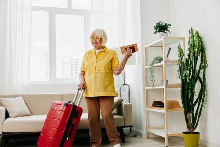 Photo for Happy senior woman with passport and travel ticket packed a red suitcase, vacation and health care. Smiling old woman joyfully stands in the house before the trip. High quality photo - Royalty Free Image