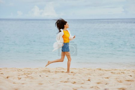 Téléchargez les photos : Femme sportive court le long de la plage en vêtements d'été sur le sable dans un T-shirt jaune et short en jean chemise blanche volant vue océan cheveux. Photo de haute qualité - en image libre de droit