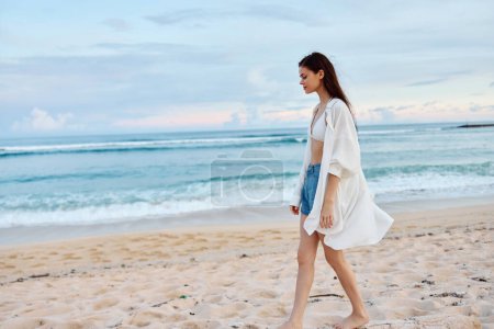 Téléchargez les photos : Joyeux bronzé femme mince en maillot de bain blanc chemise et short en jean marche sur la plage sur le sable près de l'océan avec les cheveux mouillés après la baignade, la lumière du coucher du soleil. Photo de haute qualité - en image libre de droit