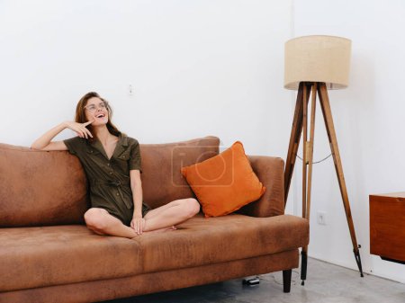 Foto de Modelo de mujer joven sentado en el sofá en casa sonriente y relajante, estilo de vida interior moderno, espacio para copiar, paleta de colores de otoño. Foto de alta calidad - Imagen libre de derechos