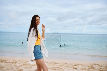Foto de Mujer deportiva corre a lo largo de la playa en ropa de verano en la arena en una camiseta sin mangas amarilla y pantalones cortos de mezclilla camisa blanca que vuela vista al mar, vacaciones en la playa y libertad de viaje. Foto de alta calidad - Imagen libre de derechos