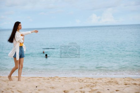 Téléchargez les photos : Femme sportive court le long de la plage en vêtements d'été sur le sable dans un débardeur jaune et short en jean chemise blanche volant cheveux vue sur l'océan, vacances à la plage et Voyage. Photo de haute qualité - en image libre de droit