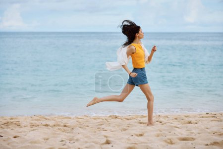 Foto de Mujer deportiva corre a lo largo de la playa en ropa de verano en la arena en una camiseta sin mangas amarilla y pantalones cortos de mezclilla camisa blanca volando vista al mar, vacaciones en la playa y viajes. Foto de alta calidad - Imagen libre de derechos