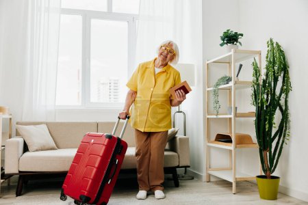 Téléchargez les photos : Heureuse femme âgée avec passeport et billet de voyage emballé une valise rouge, vacances et soins de santé. Une vieille femme souriante se tient joyeusement dans la maison avant le voyage. Photo de haute qualité - en image libre de droit
