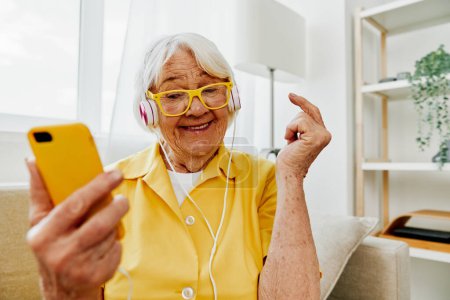 Foto de Mujer anciana feliz viendo videollamada en el teléfono y escuchando música en los auriculares, sorpresa y boca abierta, interior moderno brillante, estilo de vida de comunicación en línea. Foto de alta calidad - Imagen libre de derechos