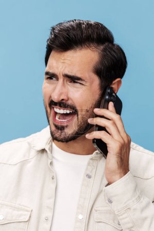 Téléchargez les photos : Portrait de colère brune masculine parlant au téléphone en gros plan, mécontent, sur un fond bleu dans un T-shirt blanc et un jean, espace de copie. Photo de haute qualité - en image libre de droit