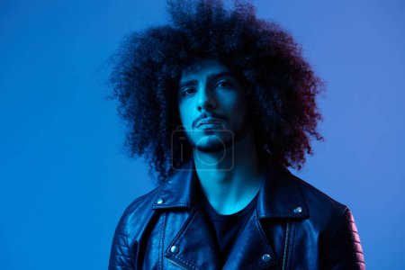 Photo for Portrait of fashion man with curly hair on blue background multinational, colored light, black leather jacket trend, modern concept. High quality photo - Royalty Free Image